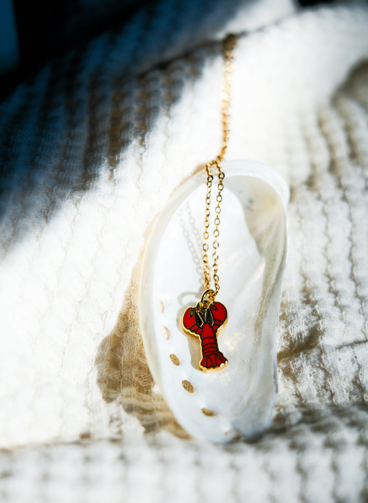 A gold chain necklace with a red lobster-shaped pendant, resting on a white seashell against a textured white knit blanket in bright natural light.