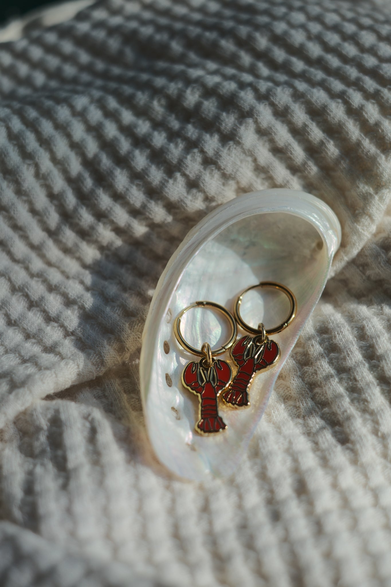 A pair of lobster-shaped keychains with red and black enamel detailing and gold-tone rings, displayed inside a white mother-of-pearl clam shell on a textured cream knit blanket.