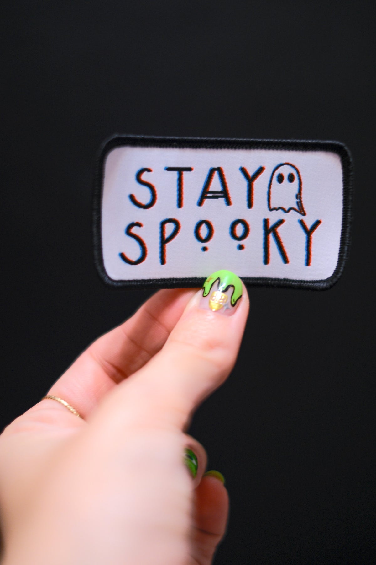Hand holding a white Stay Spooky patch with black border and ghost illustration against a dark background.