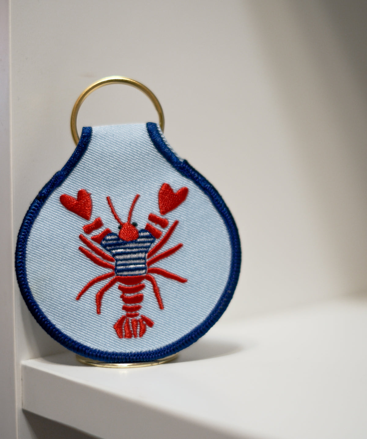 Light blue embroidered keychain featuring a red lobster in a navy striped sweater, with navy edging and a gold key ring.