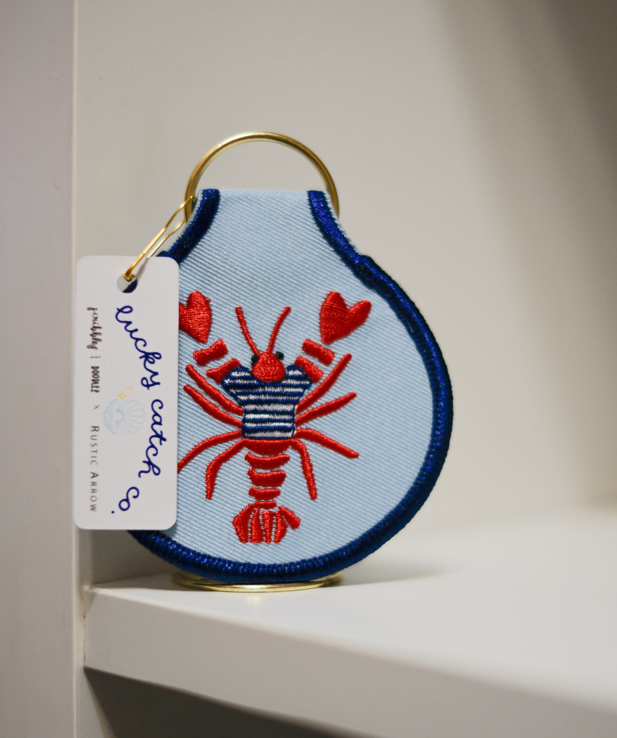Red lobster in a striped sweater embroidered on a light blue keychain with navy edging, a gold key ring, and a Lucky Catch Co. tag.