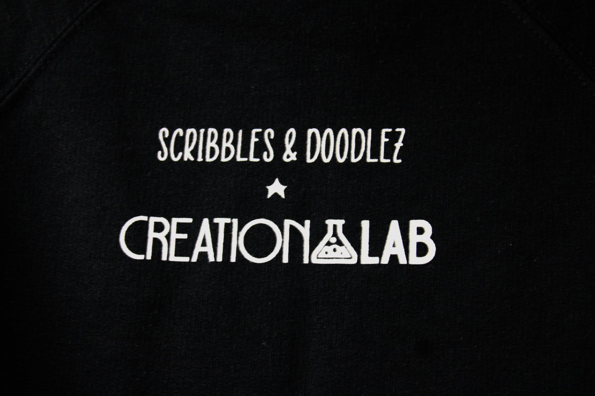 Close-up of a black garment featuring white printed text that reads 'SCRIBBLES & DOODLEZ' and 'CREATION LAB' with a small star and flask icon.