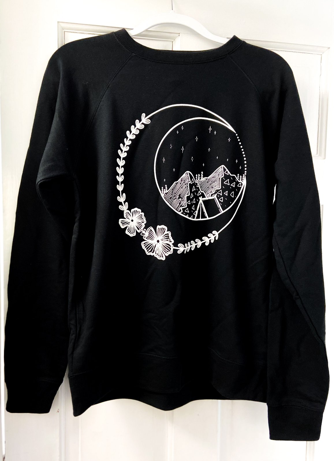 A black sweatshirt with a white design line work design, featuring a crescent moon, flowers, mountains, and a tent.