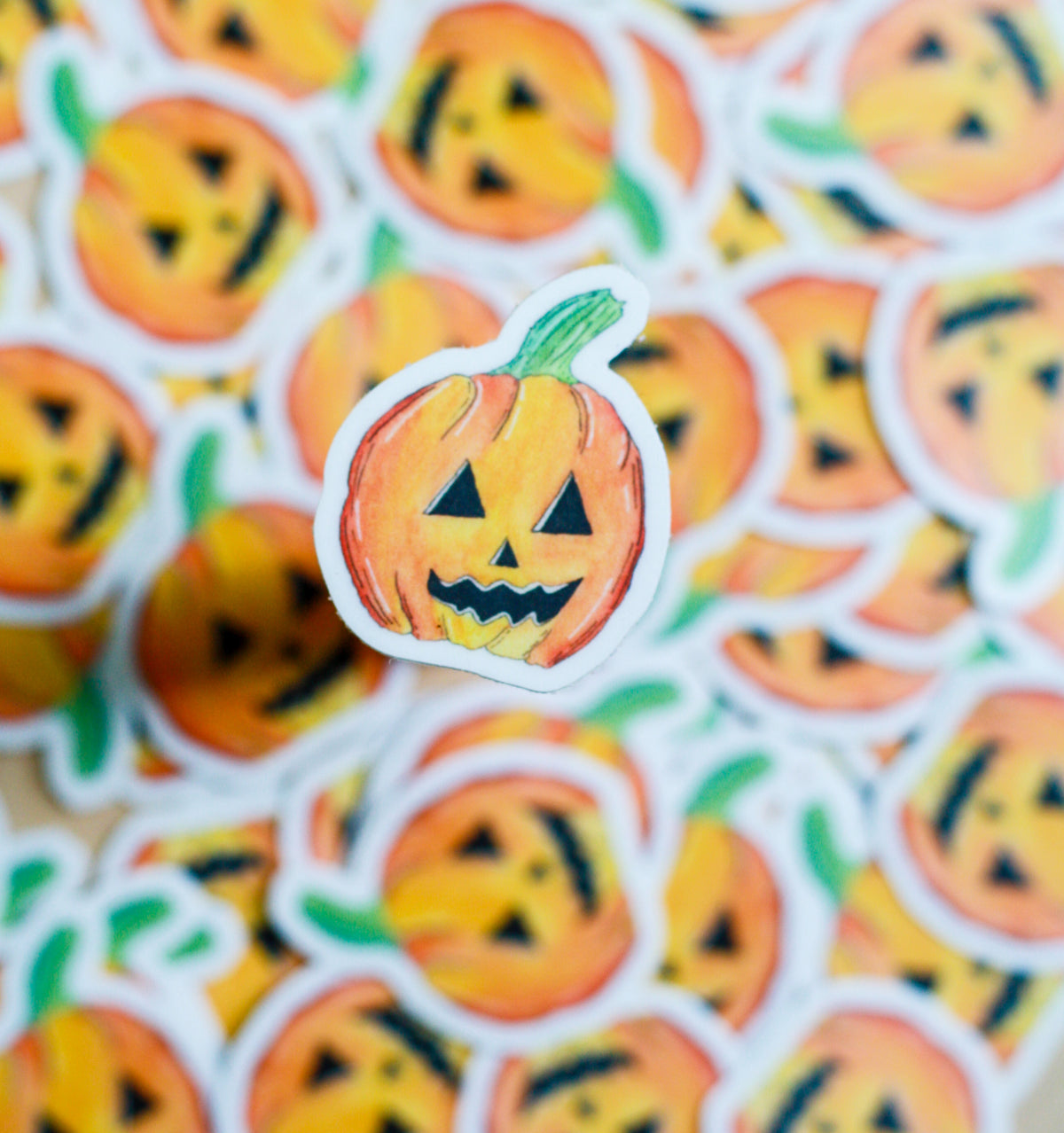 A single die-cut sticker featuring a hand-drawn orange jack-o'-lantern pumpkin with a green stem, shown in sharp focus against a blurred background of identical stickers.