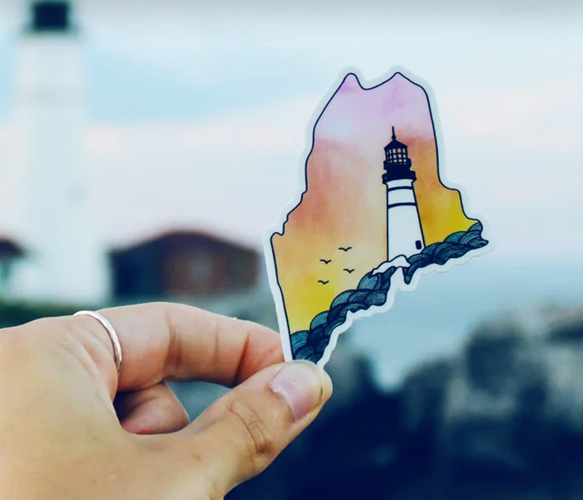 A hand holds a sticker shaped like a state outline with a lighthouse design over a sunset background.