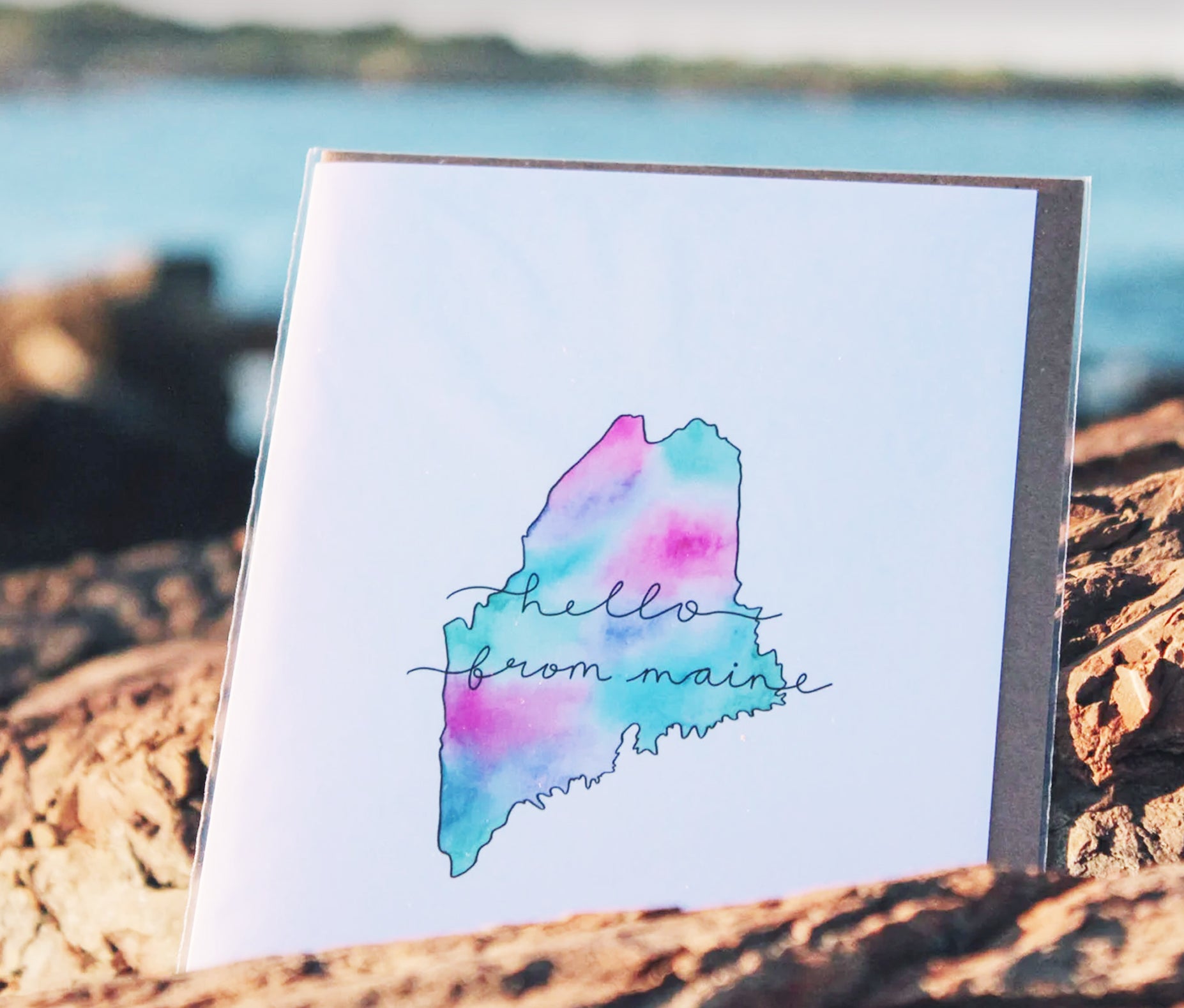 A greeting card with a watercolor outline of Maine on the cover, featuring the words hello from maine in cursive. The card is propped on rocks, with a blurred background of water and shoreline.