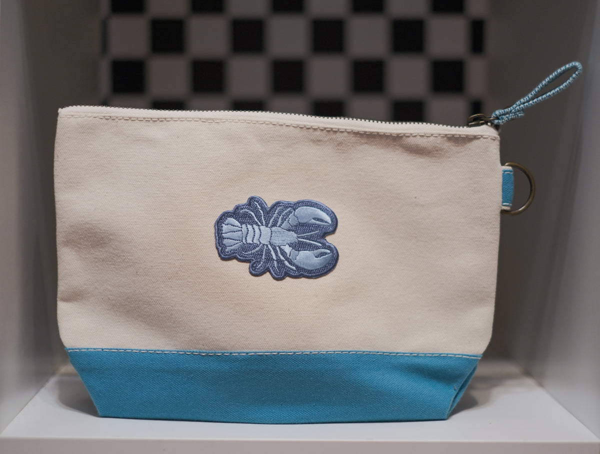 A canvas zip pouch with a cream-colored upper section, a bright blue lower panel, and a blue embroidered lobster patch on the front. It features a matching blue zipper pull.