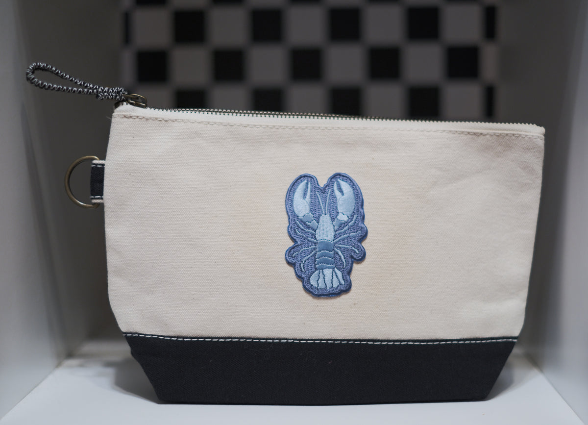 A canvas zippered pouch with a cream upper section, a black bottom panel, and a blue embroidered lobster patch in the center. It features a silver metal ring and a silver zipper pull.