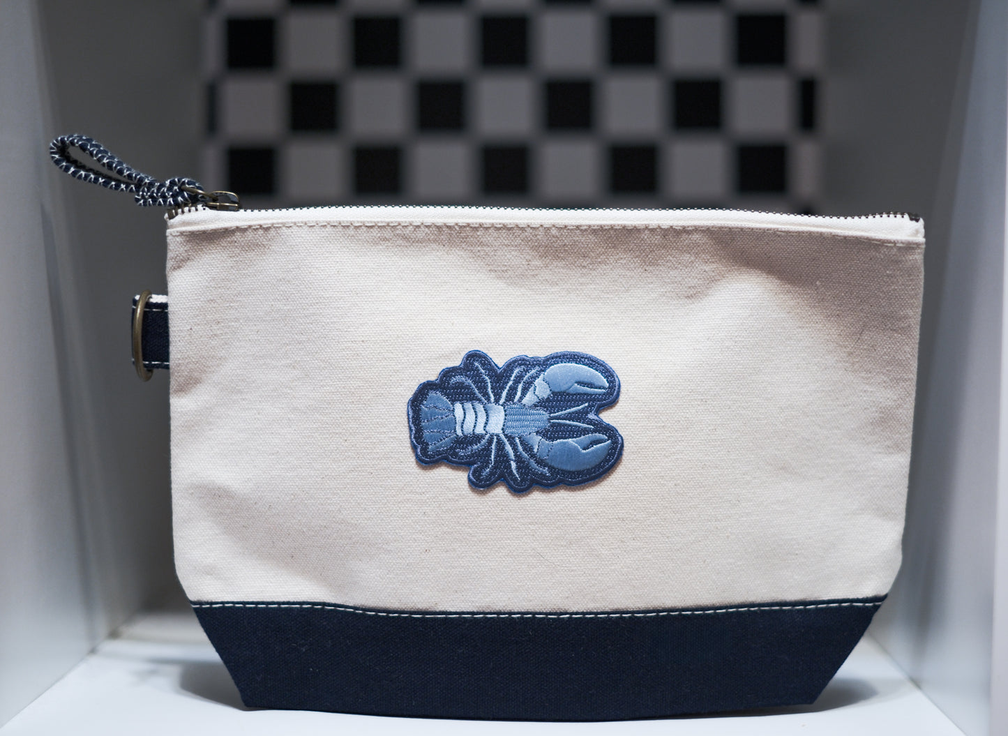 A canvas zippered pouch with an off-white top section, a dark navy bottom panel, and a blue embroidered lobster patch in the center. 