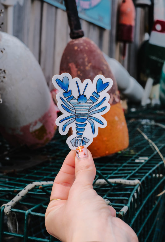 Hand holding a blue and white lobster sticker with fishing equipment in the background