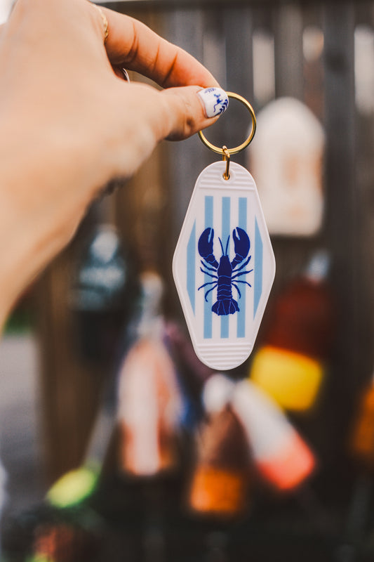 Keychain with blue lobster illustration, background of keychain blue and white stripes. Held by a hand against a blurred background.
