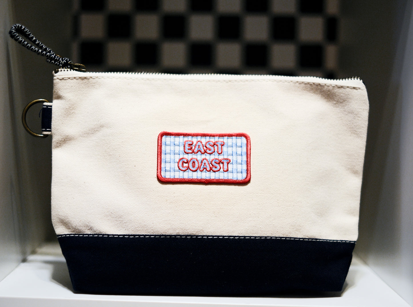 A canvas zippered pouch with a cream upper section, a navy blue bottom panel, and a central patch reading 'EAST COAST' on a red and white gingham background. A black chain strap is attached, with metal d ring.