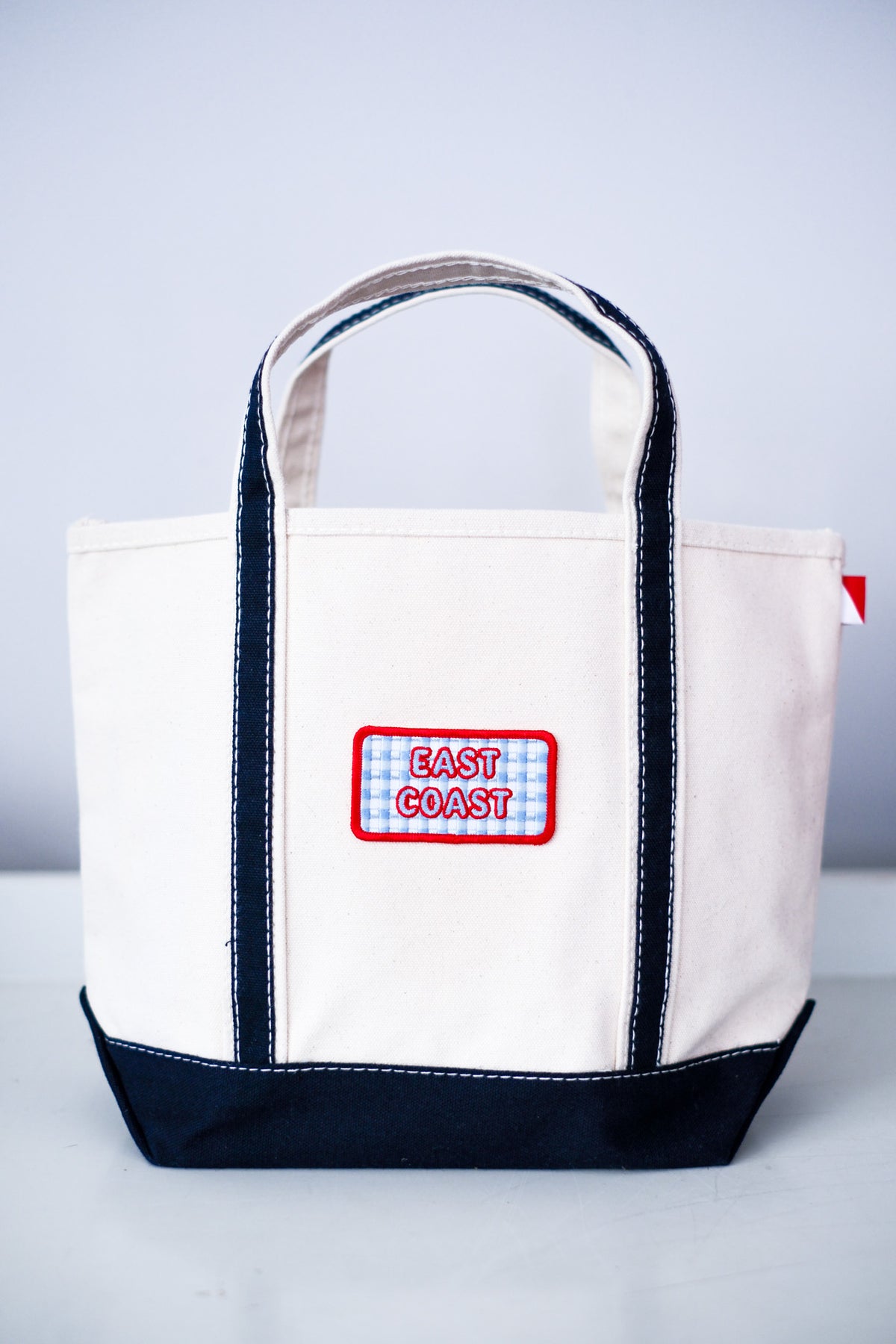A canvas tote bag with an off-white body, dark navy blue trim and bottom base, and a central patch reading 'EAST COAST' in red on a blue-and-white gingham background.
