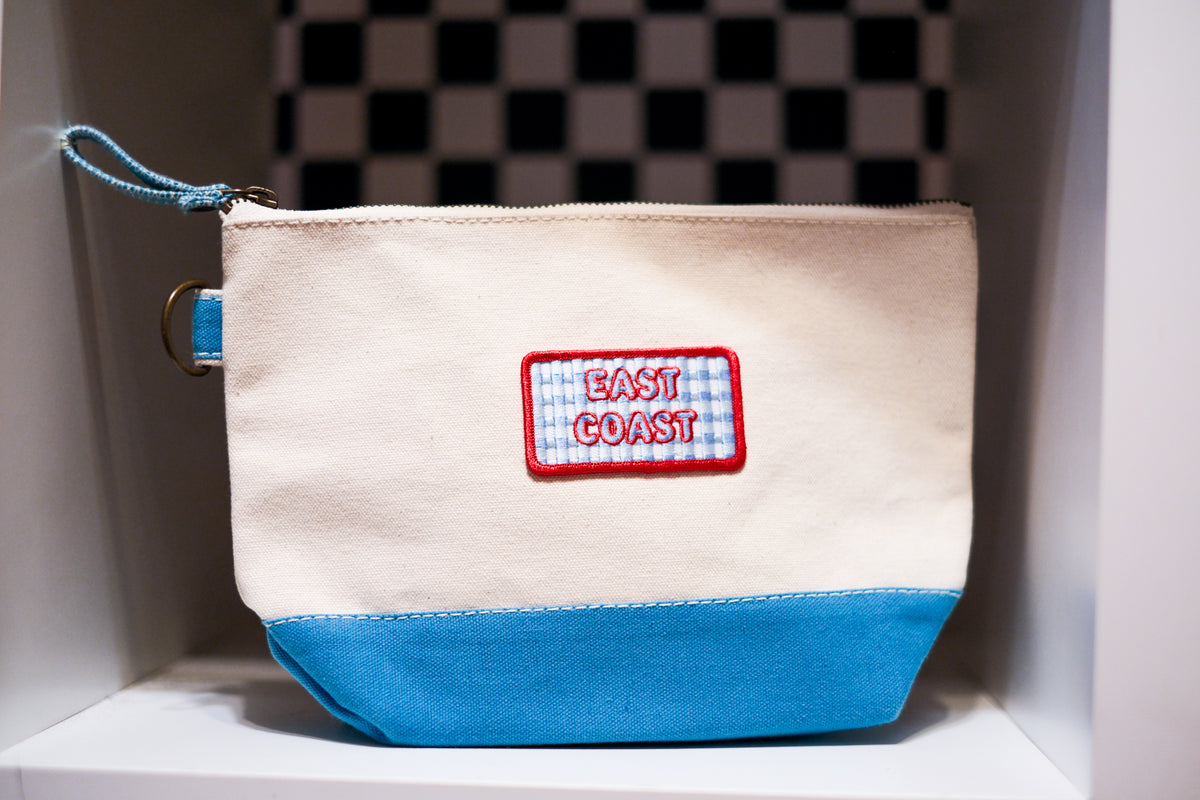 A canvas pouch with a cream upper section, a bright blue bottom panel, and a blue-and-white gingham patch on the front that reads 'EAST COAST' in red letters. The pouch has a zipper closure with a blue zipper pull and metal d ring.
