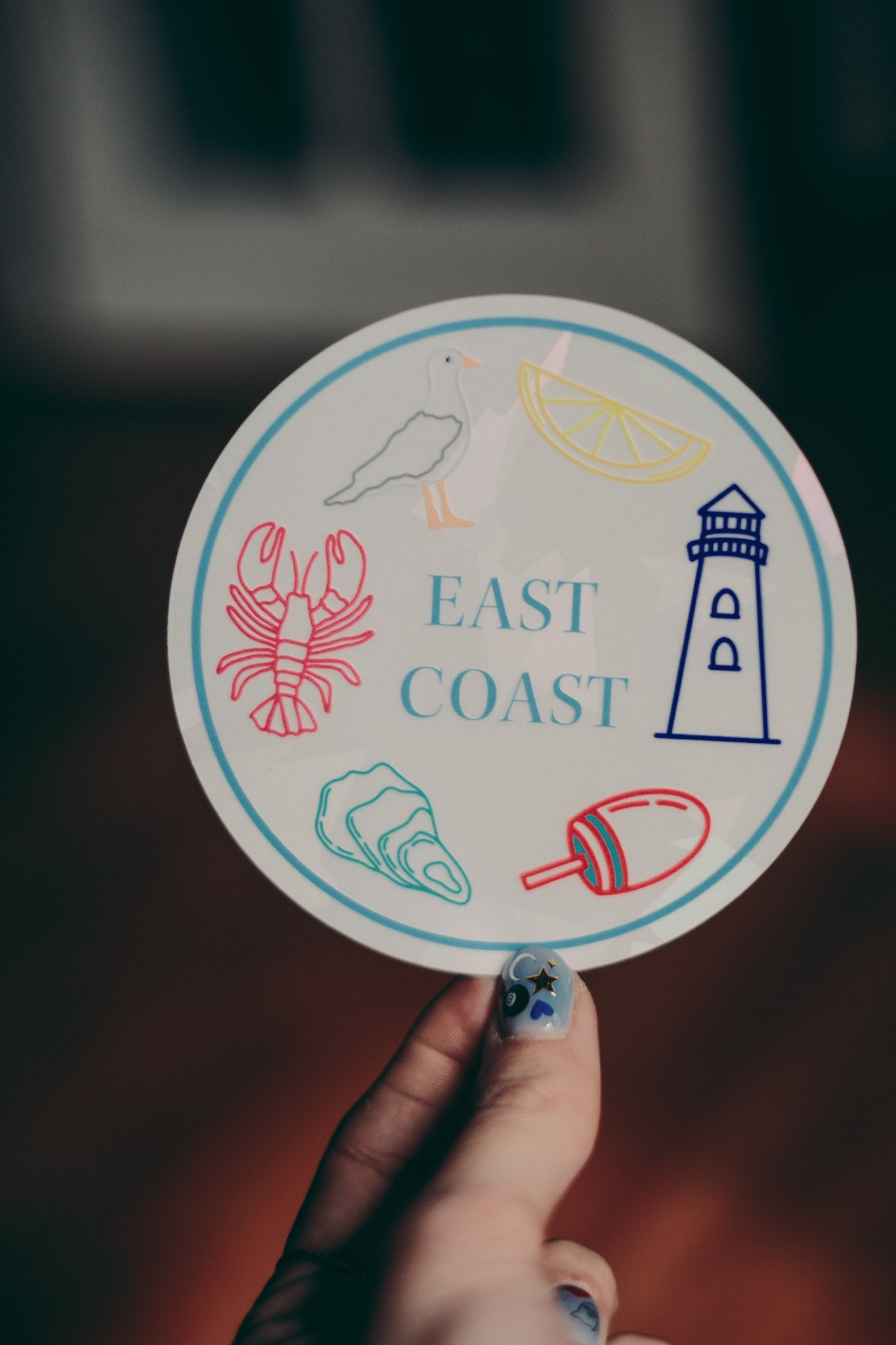 Circular sticker with coastal icons and 'East Coast' text held by a hand.