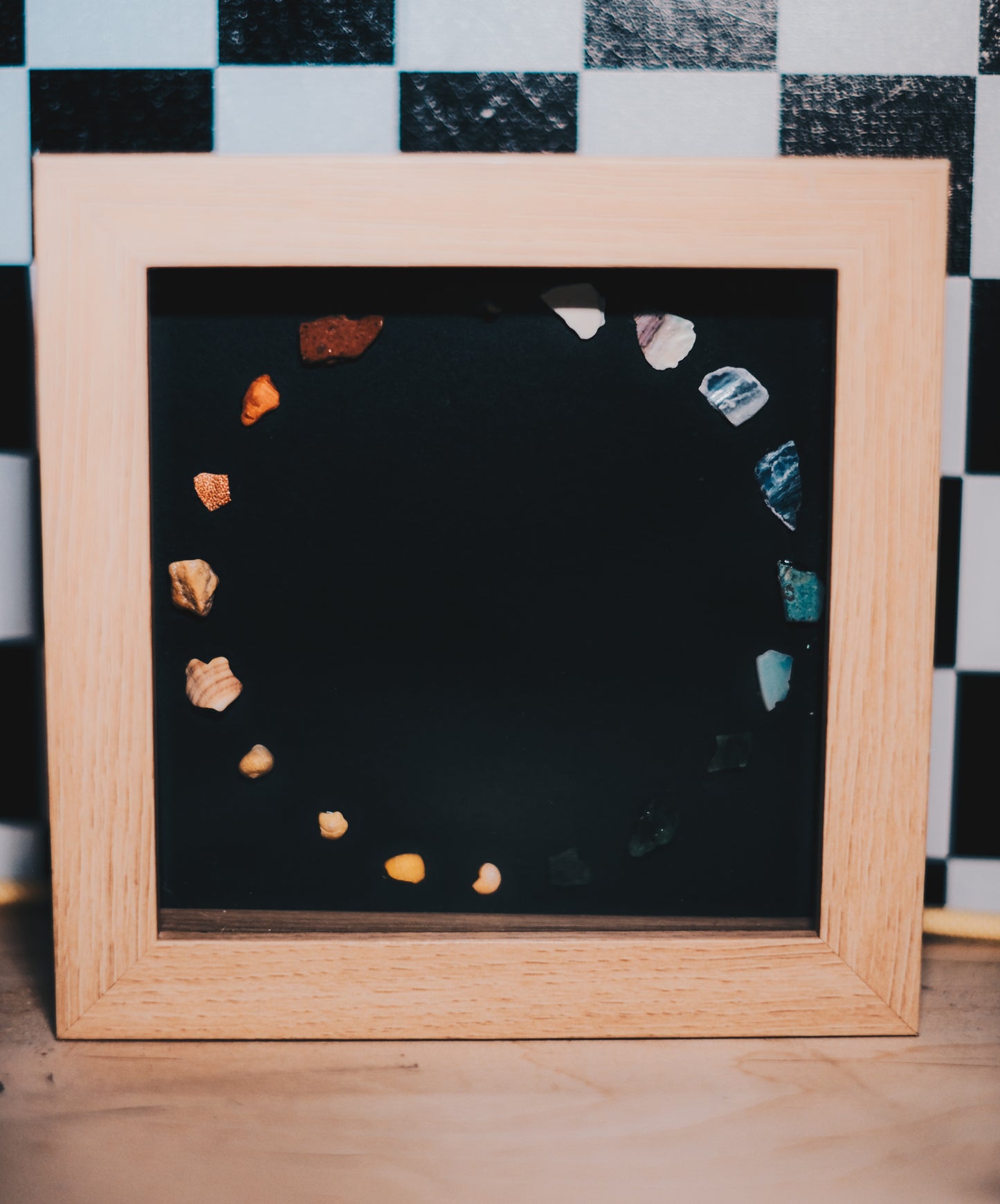Framed black board with colorful stones arranged in a circular pattern on a checkered background.