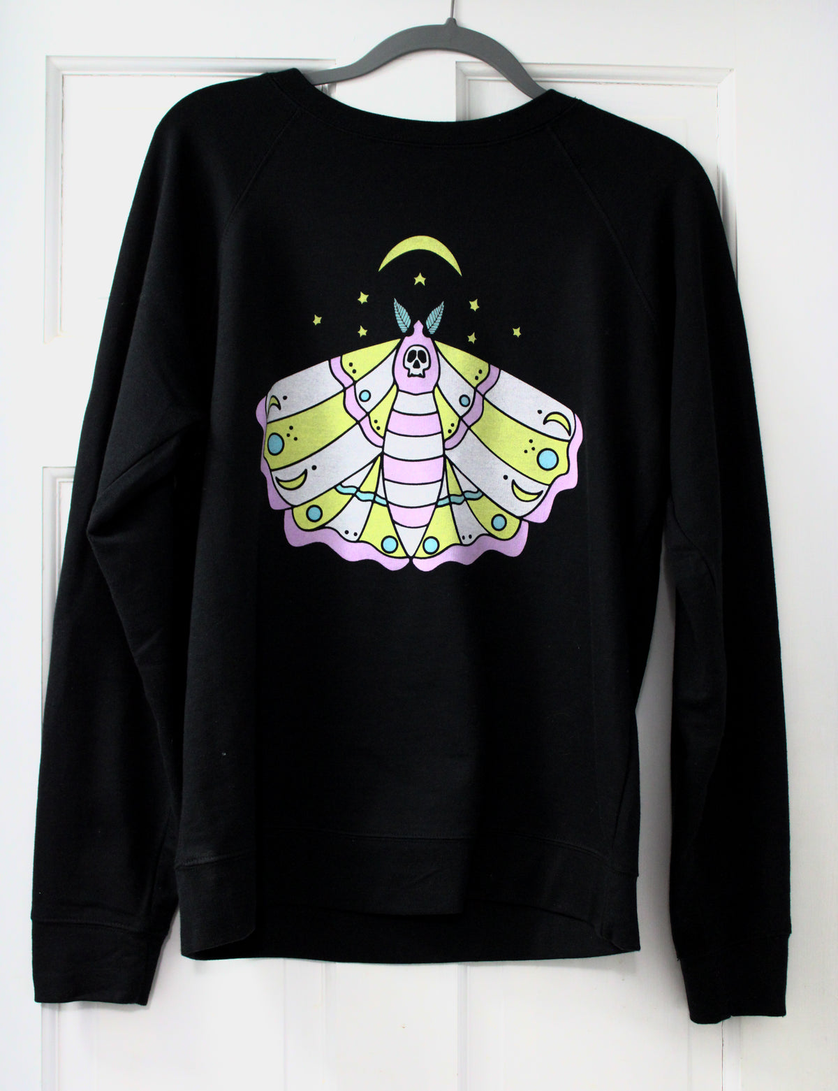 A black crewneck sweatshirt hanging on a gray hanger against a white paneled door, featuring a colorful graphic of a stylized moth with a crescent moon and stars on the chest.