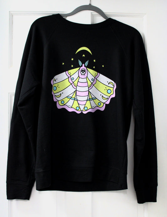 A black crewneck sweatshirt hanging on a gray hanger against a white paneled door, featuring a colorful graphic of a stylized moth with a crescent moon and stars on the chest.