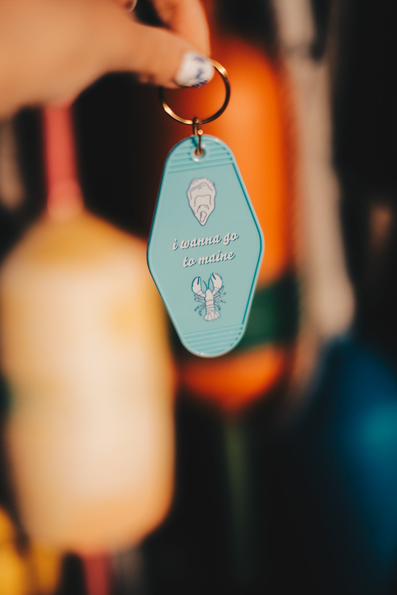 Keychain with a blue tag and lobster illustration, shell illustration, and the text "i wanna go to maine" held by a hand against a blurred background.