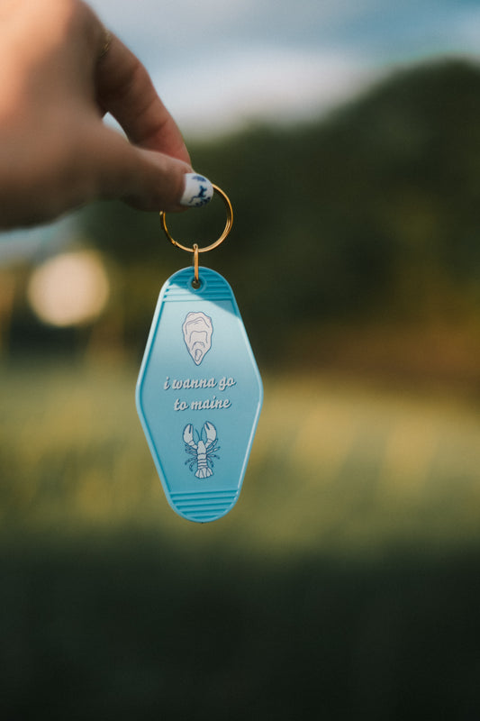 Blue keychain tag with "I wanna go to maine" with shell and lobster graphic. Held by a hand against a blurred natural background.