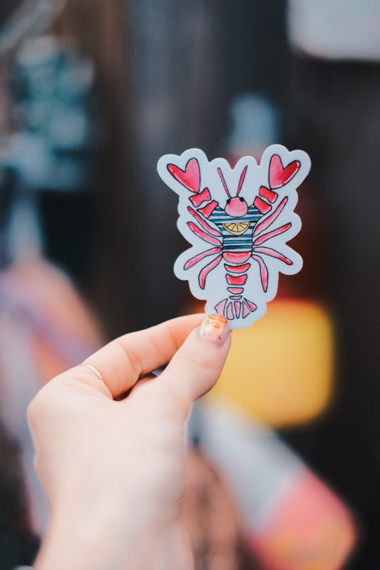 Hand holding a colorful sticker of a lobster with hearts on a blurred background