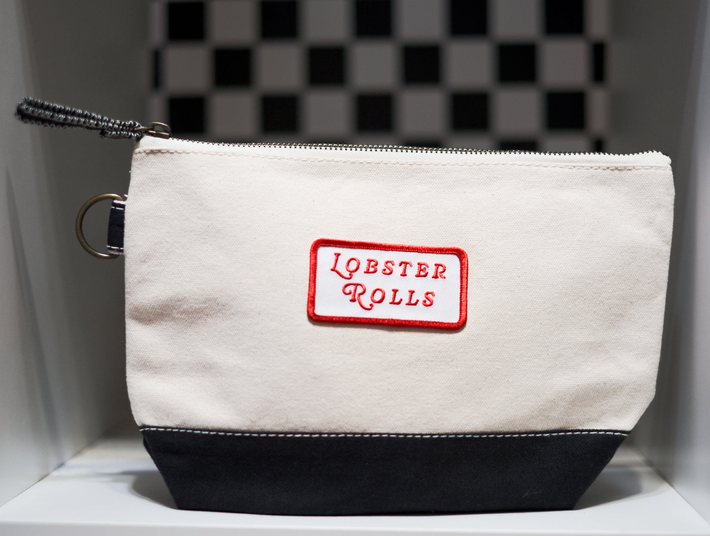 A canvas zip pouch with an off-white upper section, a black bottom panel, and a red embroidered patch reading 'LOBSTER ROLLS' on the front. 