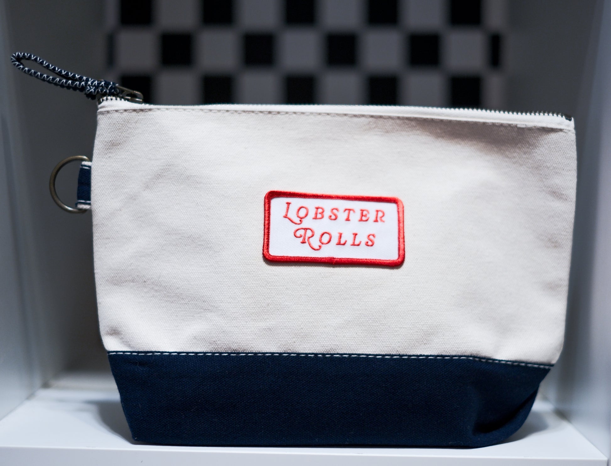 A canvas zippered pouch with an off-white upper section, a navy blue bottom panel, and a red rectangular patch reading 'LOBSTER ROLLS' on the front. 