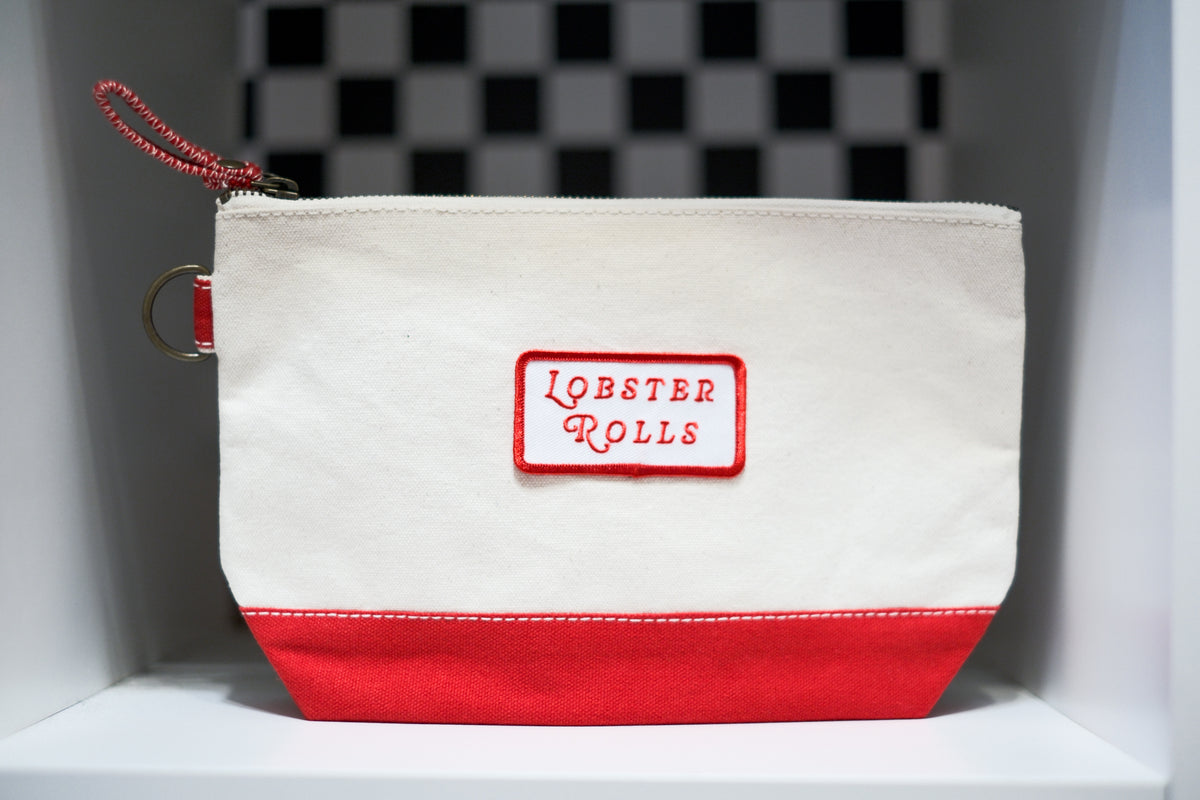 A canvas zippered pouch with an off-white top and a red bottom panel, featuring a red rectangular patch with the words 'LOBSTER ROLLS' in red script. A red zipper pull and metal ring are attached on the left side.