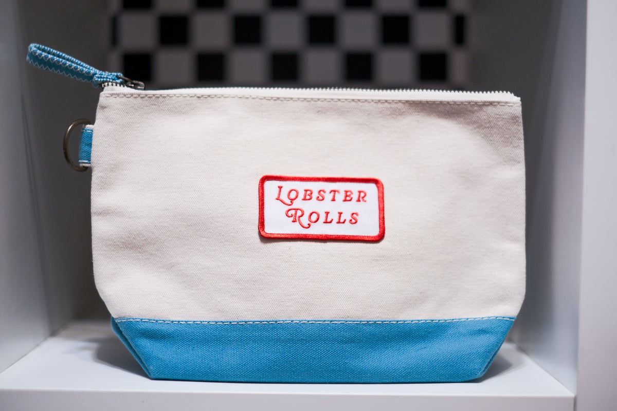 A canvas zipper pouch with a cream body, a light blue bottom panel, and a blue zipper pull. A red-bordered patch on the front reads 'LOBSTER ROLLS' in red lettering.