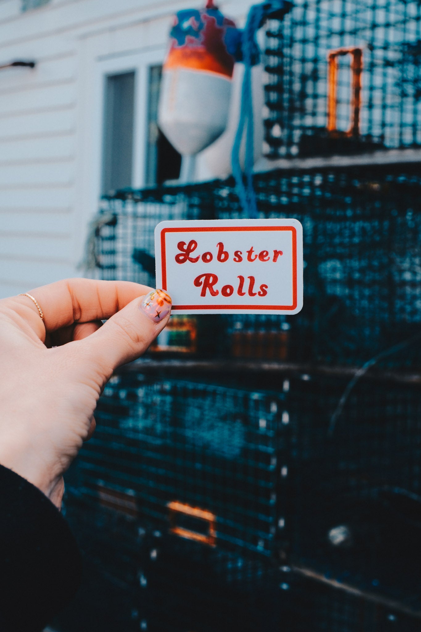 Hand holding a 'Lobster Rolls' sticker in front of lobster traps.