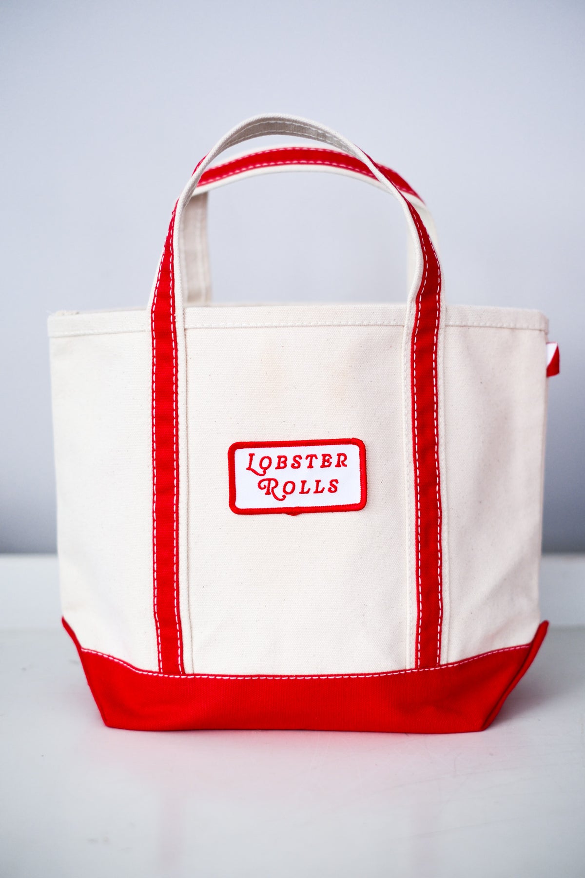 A canvas tote bag in off-white with red trim, red handles, and a red bottom panel. A front patch reads 'LOBSTER ROLLS' in red text on a white background.
