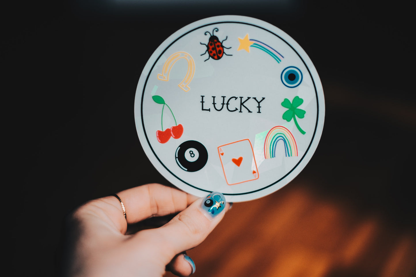Round decal with various good luck symbols and the word 'LUCKY' held by a hand against a dark background