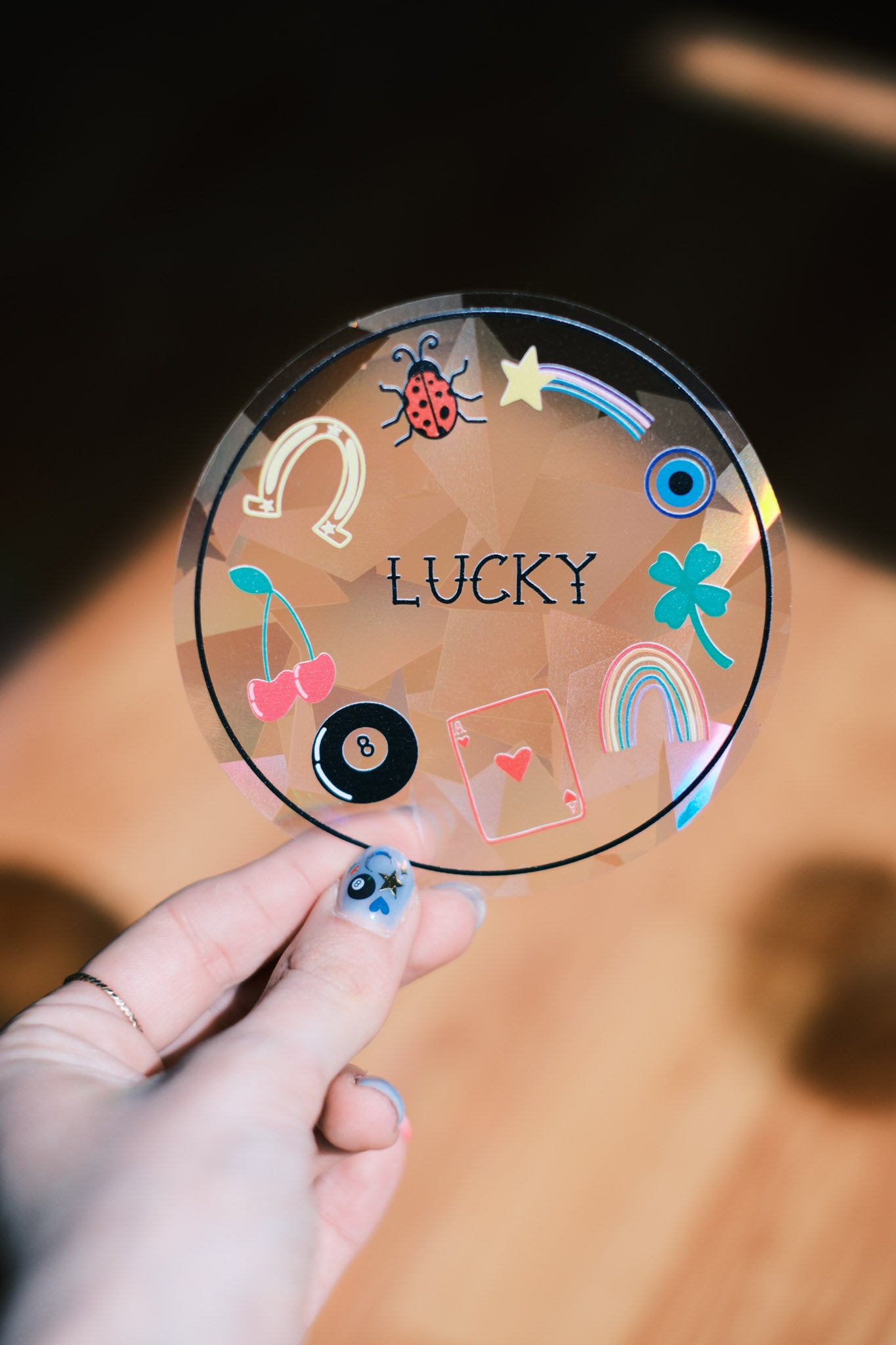 Round transparent sticker with colorful good luck symbols and the word 'LUCKY' held by a hand.
