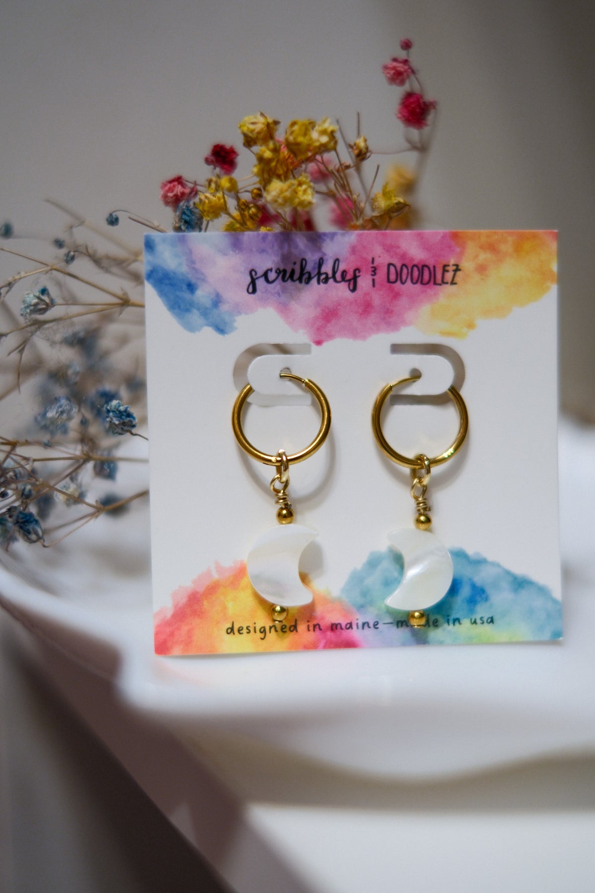 Gold hoop earrings with white crescent moon pendants, displayed on a colorful card that reads 'scribbles + doodlez', with dried flowers in the background.