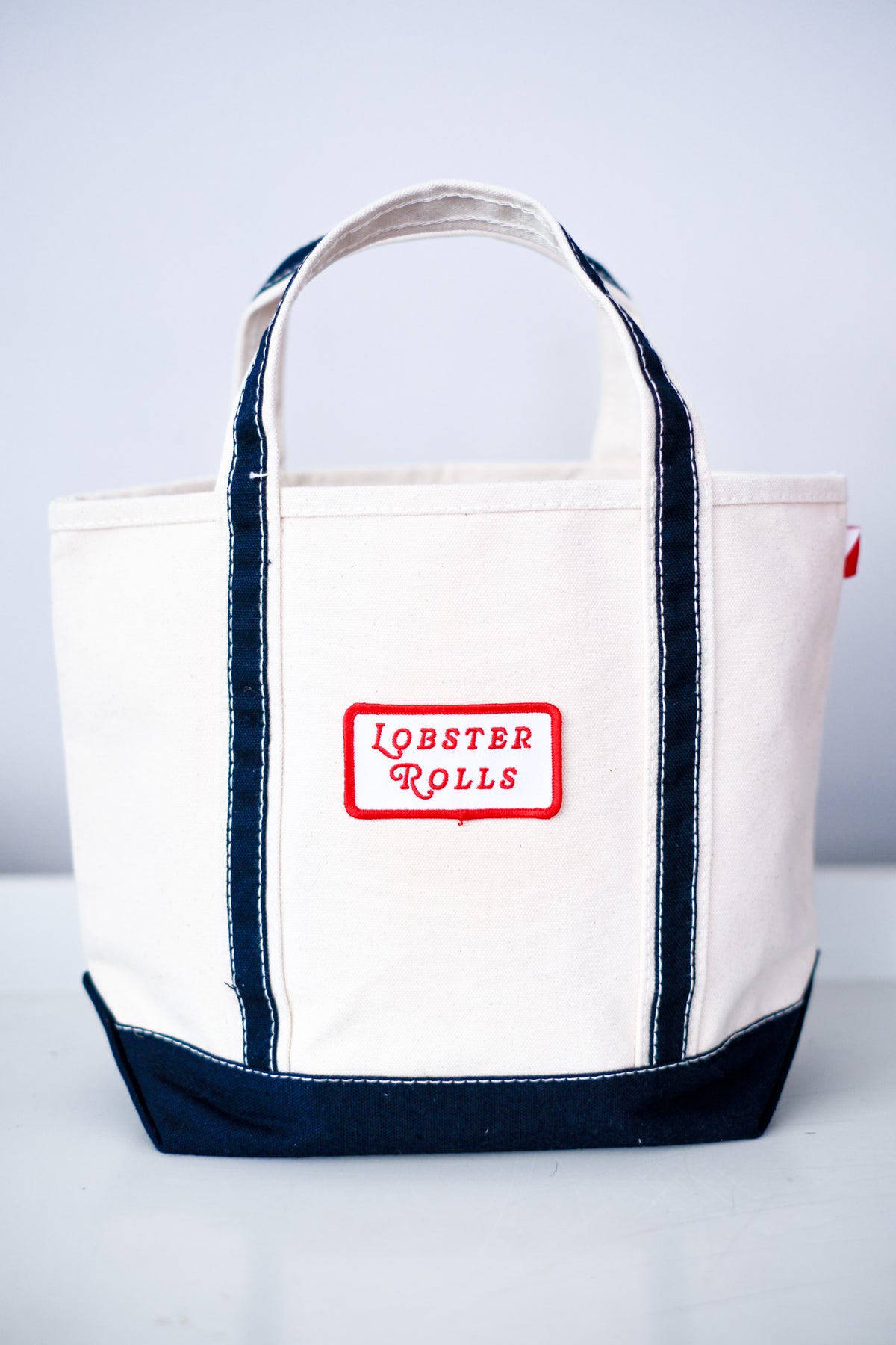 A canvas tote bag with a cream body, dark navy blue trim and bottom, and a red embroidered patch on the front that reads 'LOBSTER ROLLS'.