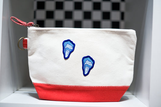 A canvas zippered pouch with a white upper section, a red bottom section with two blue and white oyster patches on the front. On the left side a red zipper pull and a metal d-ring.