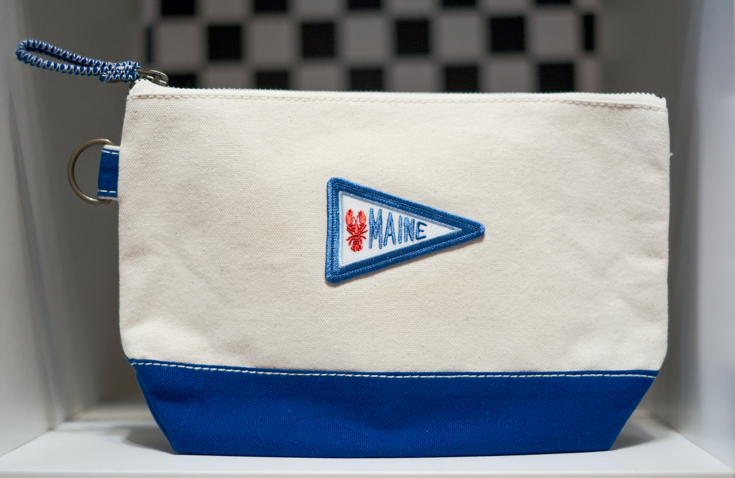 A canvas zippered pouch with a cream upper section and a royal blue lower section, featuring a blue, white, and red embroidered 'MAINE' lobster pennant on the front. It has a blue braided zipper pull and a metal ring attachment.