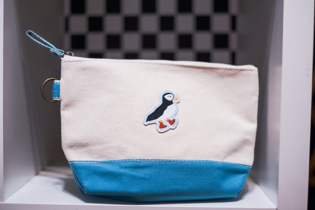 A canvas zip pouch with a cream-colored top and a bright blue bottom panel, featuring a puffin patch on the front and a matching blue zipper pull.