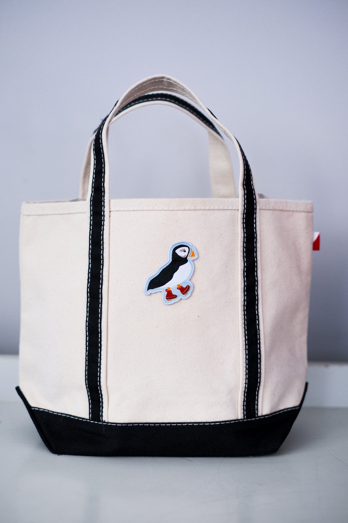An off-white canvas tote bag with black trim, black bottom panel, and a colorful puffin patch on the front, set against a neutral gray background.