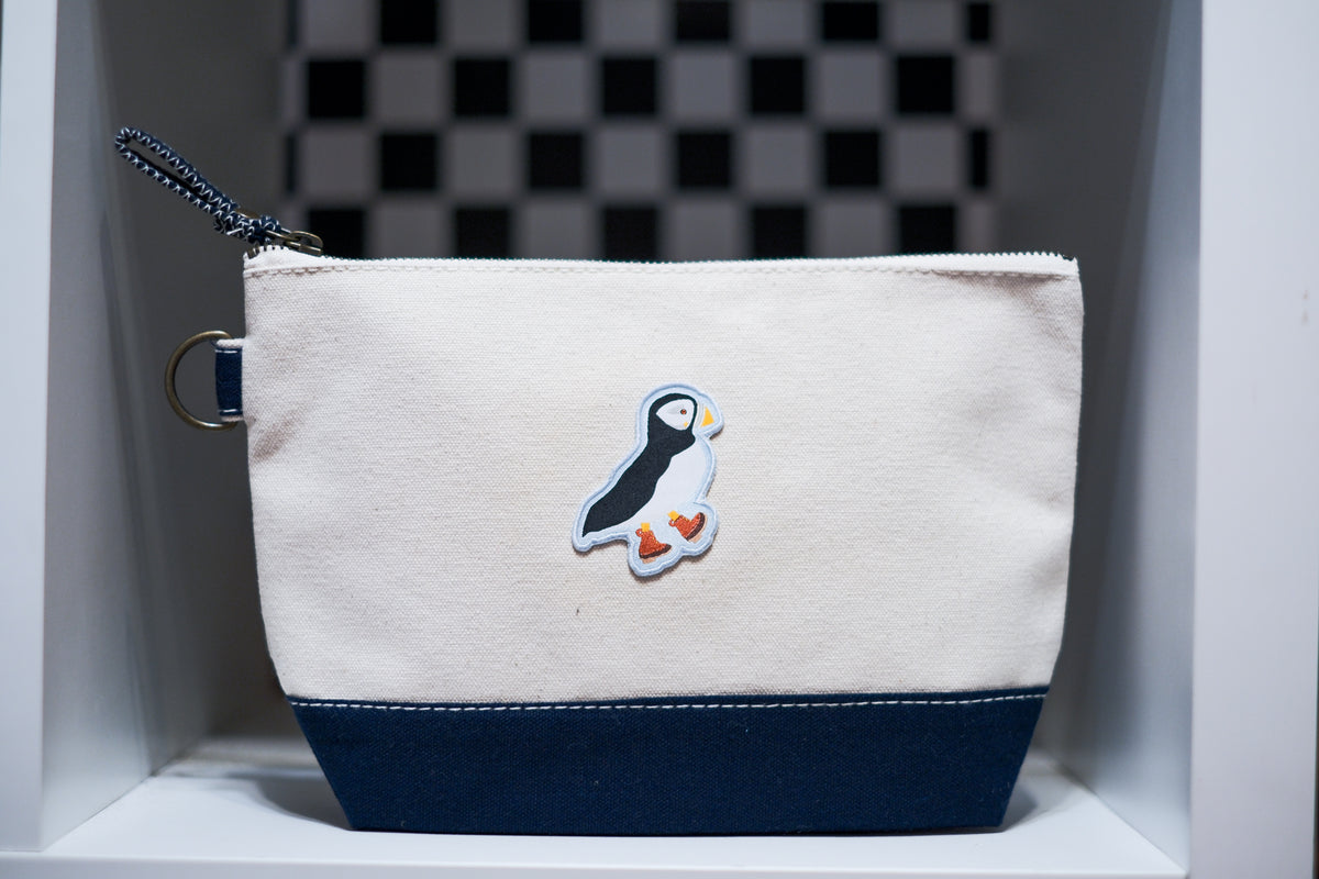 A canvas zippered pouch with an off-white top half, a navy blue bottom panel, and a puffin patch on the front centered.
