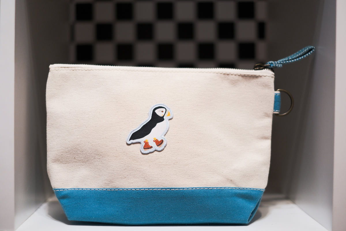 A canvas zip pouch with a cream-colored top and a bright blue bottom panel, featuring a puffin patch on the front and a matching blue zipper pull.
