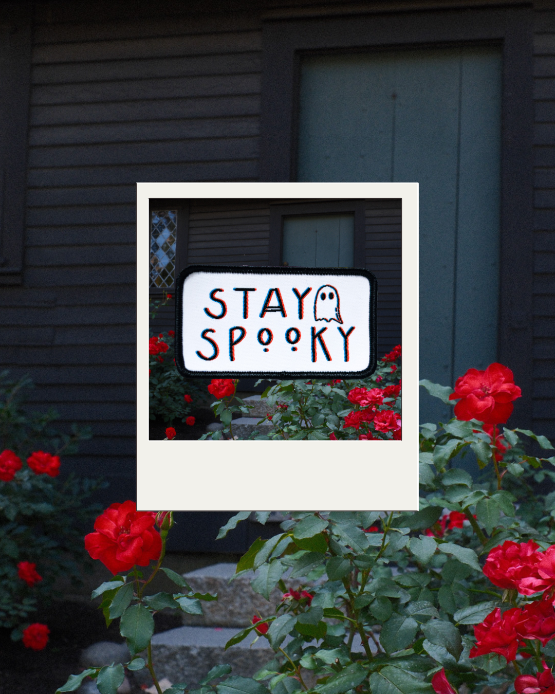 White Stay Spooky patch with black border and ghost illustration, framed over red roses outside a dark house.