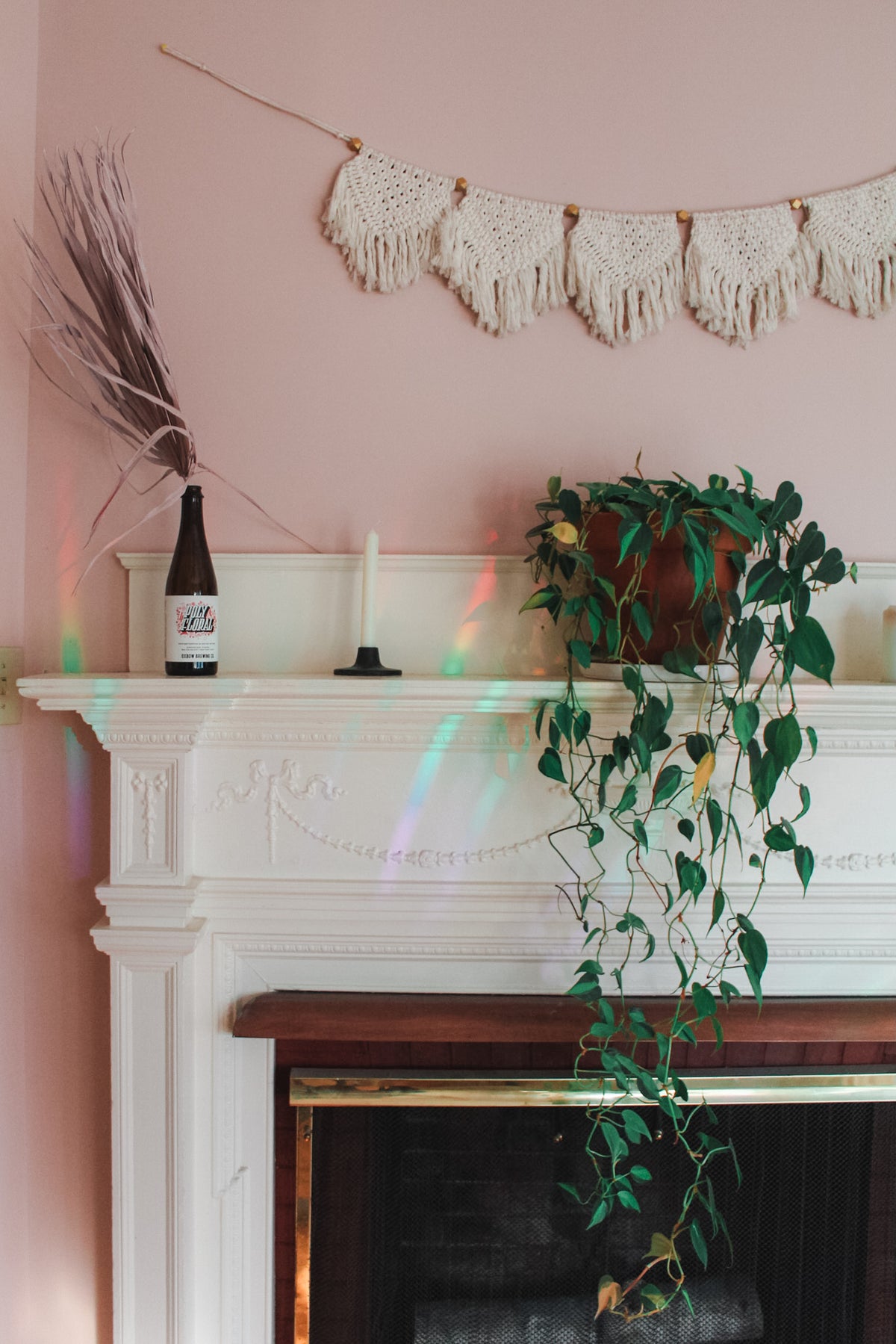 A white fireplace mantel in a pink room decorated with a cream macrame banner, a green trailing plant, a dried grass stem, and a dark bottle, and rainbow beams.